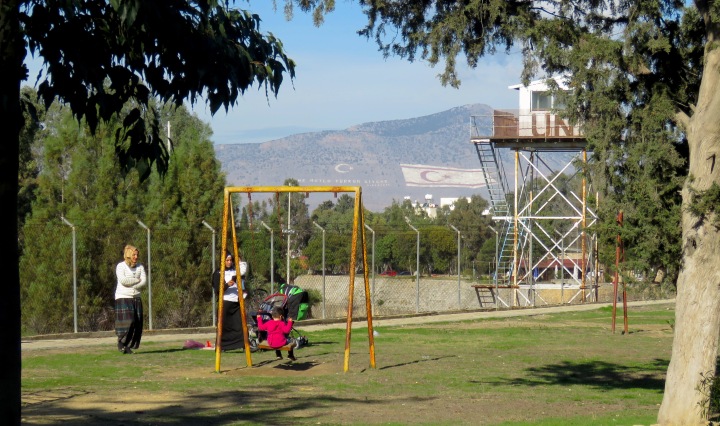 Two Turkish Cypriot Women and their children play in Nicosia's Forbidden Zone with two stark reminders of the occupation on nearby mountains - by Anika Mikkelson - Miss Maps - www.MissMaps.com